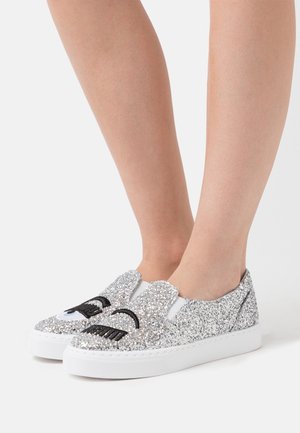 Zapatillas slip-on plateadas con purpurina y suela blanca, que presentan ojos y bigotes al estilo de dibujo animado en negro y azul en la parte frontal.