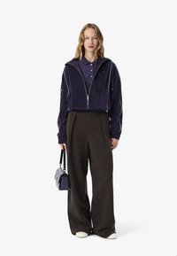 Giacca corta in velluto viola con piping bianco, abbinata a pantaloni larghi marrone scuro e una piccola borsa nera. Il modello indossa una polo viola.