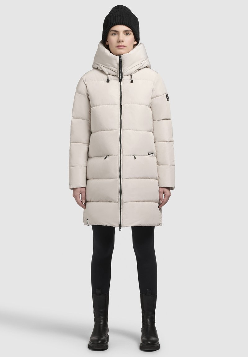 Beige pufferjack met een hoge kraag en capuchon, voorzien van een rits aan de voorkant, een diagonaal gestikt patroon en zijzakken. Draag het over zwarte legging en laarzen.