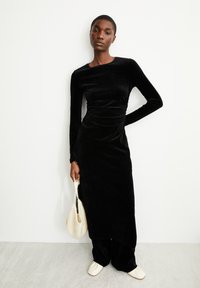 Schwarzes Samtkleid mit langen Ärmeln, tailliertem Oberteil und drapierten Details, kombiniert mit einer cremefarbenen Stricktasche und weichen flachen Schuhen.