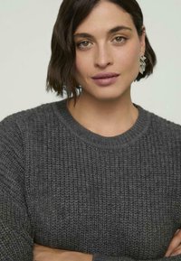 Szary sweter dziany z okrągłym dekoltem, teksturowym wzorem i ściągaczami. Modelka ma krótkie brązowe włosy i nosi srebrne kolczyki w kształcie liści.