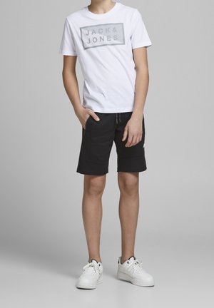 T-shirt blanc avec logo gris "JACK & JONES", associé à un short noir avec fermetures éclair latérales. Des baskets blanches complètent la tenue.