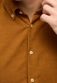 Camicia in velluto a coste marrone con colletto button-down, bottoni bianchi, texture visibile e dettaglio di pattina ordinata.