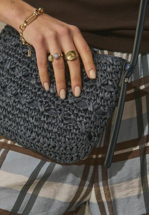 Ręka z złotymi pierścieniami i bransoletką trzyma czarną plecioną torebkę na tle spodni w kratę i brązowego topu.