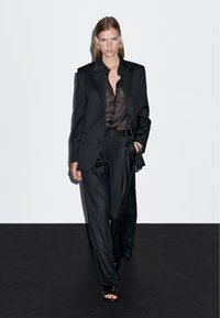 Czarny elegancki garnitur z wyrafinowaną marynarką i luźnymi spodniami, zestawiony z ciemną półprzezroczystą bluzką i czarnymi sandałami na paski.