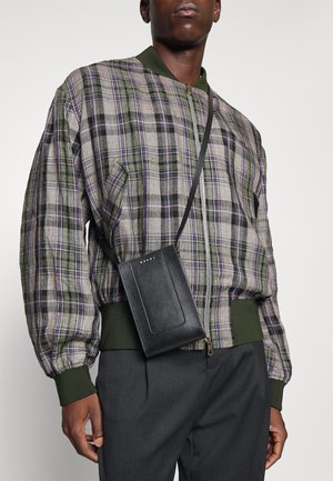 Giubbotto bomber a quadri verde e grigio con dettagli viola, abbinato a una piccola borsa a tracolla in pelle nera con una texture liscia e un design minimale.