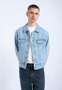 Chaqueta de mezclilla azul claro con dos bolsillos en el pecho, botones plateados y un corte relajado, combinada con una camiseta blanca y jeans azul oscuro.