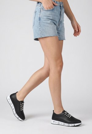 Osoba chodící, oblečená v světle modrých džínových šortkách a černých teniskách s bílými podrážkami s hvězdnými a textovými vzory.
