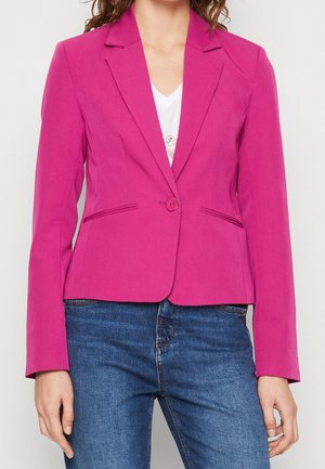 Frau trägt ein hellrosa Blazer mit einem Knopf über weißem Oberteil und blauer Jeans, steht vor weißem Hintergrund.