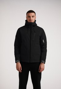 Giacca nera isolante con colletto alto, cerniera frontale e tasca sul petto con zip. Caratterizzata da dettagli con logo e accenti bianchi sulle maniche.