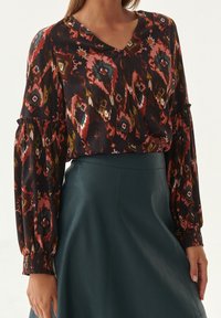 Blouse à manches longues avec un motif géométrique multicolore sur fond sombre, décolleté en V et poignets froncés. Associée à une jupe en cuir verte.