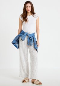 Mujer joven con top sin mangas blanco, pantalones anchos a rayas, chaqueta de mezclilla atada a la cintura y sandalias beige, de pie sobre fondo blanco.