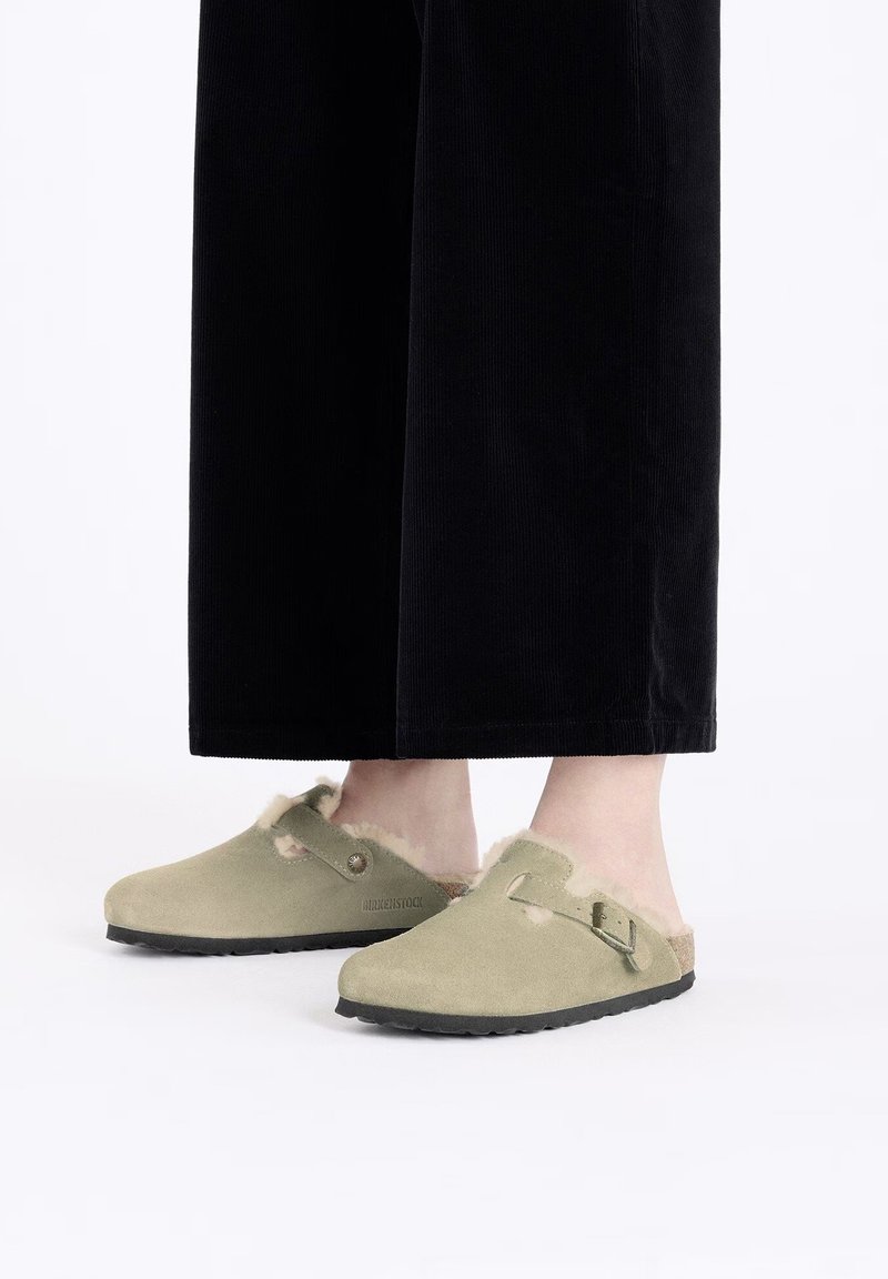 Zuecos de ante verde oliva con forro de piel sintética, correa ajustable con hebilla y suelas de goma negra. Llevados con pantalones de pana negra.