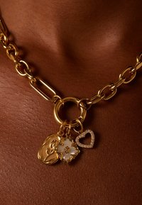 Collier en chaîne dorée avec des charms décoratifs : un visage en contour doré, une fleur blanche avec des accents dorés, et un pendentif en forme de cœur orné de strass.