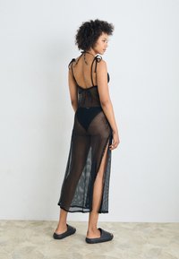 Črna mrežasta pokrivala z razporki, ki ima vezi na ramenih, v kombinaciji s črnimi bikini hlačkami. Model nosi črne natikače.