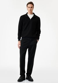 Svart ribbad tröja med dragkedja, bärs över en vit topp. Mörka byxor och svarta skor fulländar outfiten. Enkel, modern design.