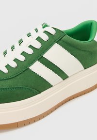 Adidași verzi din suede cu detalii albe pe dungi, șireturi albe, o talpă groasă de culoare crem și o talpă din gumă. Finisaj texturat.