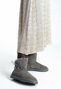 Botas de tobillo de ante gris con un puño esponjoso y un lazo decorativo, usadas con una falda crema moteada en un fondo neutro.