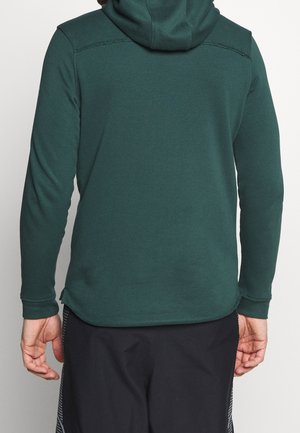 Man draagt een donkergroene hoodie met lange mouwen en zwarte sportshorts, gezien van achteren tegen een effen achtergrond.