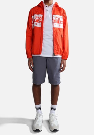 Veste légère rouge avec des détails de logo blancs, manches courtes, associée à un polo blanc, un short gris et des sneakers blanches avec des accents noirs.