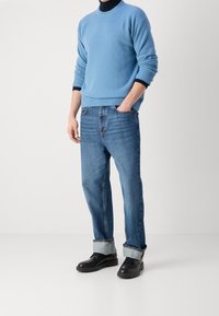 Hellblauer, gestrickter Pullover mit gerippten Bündchen, kombiniert mit dunkelblauen Jeans mit aufgekrempelten Säumen. Schwarze Schnürschuhe runden das Outfit ab.