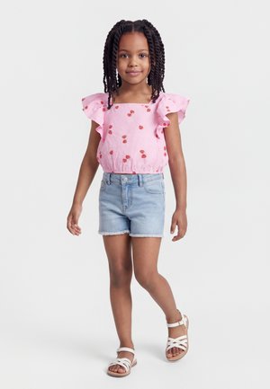 Niña con el cabello trenzado, vestida con una blusa rosa con estampado de fresas, pantalones cortos de mezclilla y sandalias blancas, posando contra un fondo liso.