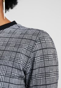 Pull en maille avec un motif à carreaux noir et blanc, présentant des lignes texturées et un col côtelé. Gros plan sur la zone de l'épaule.