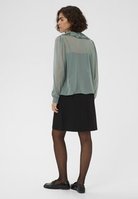 Transparante, lichtgroene blouse met gerimpelde kraag, elastische mouwen; gecombineerd met een zwarte rok en zwarte loafers, wat een minimalistische stijl accentueert.