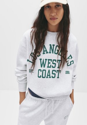 Lichtgrijze sweatshirt met een graphic "Los Angeles West Coast" in donkergroen. Voorzien van geribbelde manchetten en een ontspannen pasvorm. Gecombineerd met bijpassende grijze sweatpants.