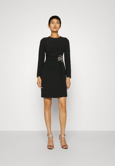 Abito nero con maniche lunghe, silhouette aderente, dettaglio arricciato sul lato e un'elegante accentatura in gioiello in vita. Tessuto dalla texture liscia.