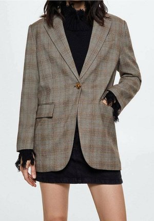 Frau trägt braunen karierten Blazer über schwarzem Pullover mit ausgefransten Kanten und schwarzem Rock, eine Hand in der Blasertasche.