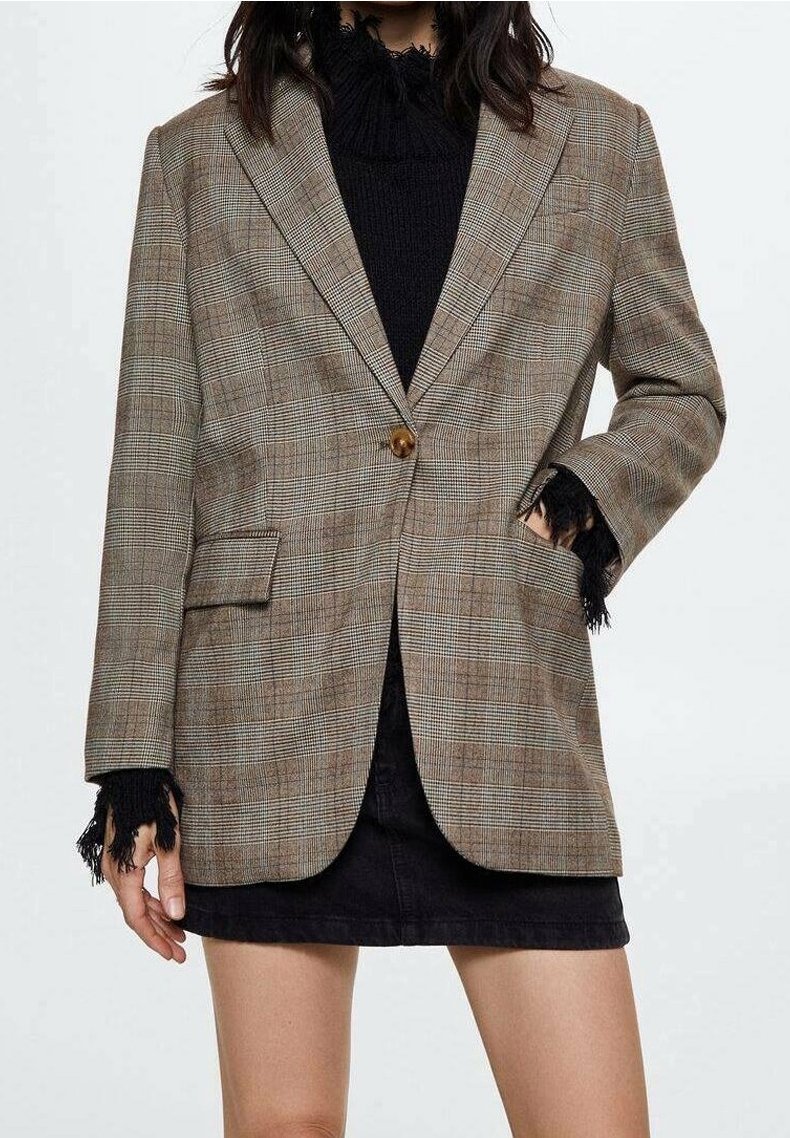 Femme portant un blazer à carreaux marron sur un pull noir aux bords effilochés et une jupe noire, une main dans la poche du blazer.