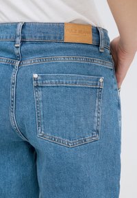 Vaqueros de mezclilla azul con una etiqueta de cuero marrón en la parte trasera que dice "PULZ JEANS", con bolsillos cosidos y detalles de herrajes visibles.