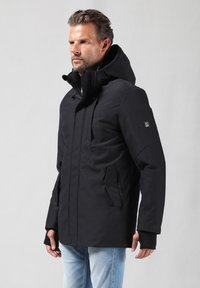 Chaqueta negra con capucha y cierre frontal, dos bolsillos laterales, tejido texturizado y silueta ajustada, usada sobre jeans azul claro.