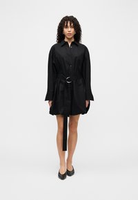 Μαύρο φόρεμα πουκάμισο με μακριά μανίκια, κούμπωμα με κουμπιά και ζώνη στη μέση με ασημένιο κρίκο. Λείο ύφασμα, μήκος πάνω από το γόνατο. Απλός σχεδιασμός.