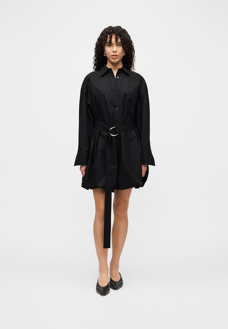 Μαύρο φόρεμα πουκάμισο με μακριά μανίκια, κούμπωμα με κουμπιά και ζώνη στη μέση με ασημένιο κρίκο. Λείο ύφασμα, μήκος πάνω από το γόνατο. Απλός σχεδιασμός.