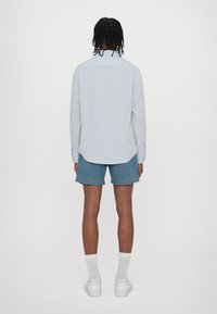 Světlomodrá košile s dlouhým rukávem, zadním klínem a zaobleným lemem, v kombinaci s džínovými šortkami. Celý outfit doplňují bílé kotníkové ponožky a tenisky.