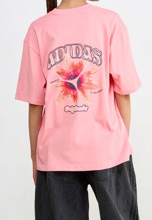 Osoba nosząca luźną różową koszulkę Adidas z dużą czerwoną i pomarańczową grafiką kwiatu oraz tekstem na plecach, w połączeniu z ciemnymi dżinsami.