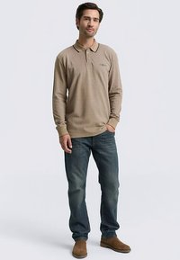 Pikkade varrukatega beež polo-särk kontrastse kraega, siniste teksade ja pruunide seemisnahast saapadega. Modell seisab ühevärvilise tausta ees.