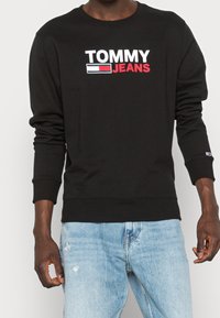 Sudadera negra con el logo "TOMMY JEANS" en blanco y rojo. Puños y dobladillo acanalados. Combinada con jeans claros de lavado desgastado.