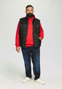 Schwarweste mit Füllung über einem roten Pullover, blauen Jeans und weißen Sneakers. Die Weste verfügt über einen Reißverschluss und Taschen mit einem Logo auf der Brust.