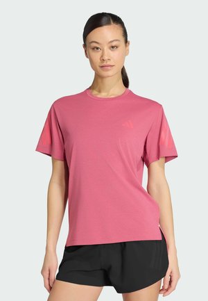 Donna con capelli scuri raccolti in una coda di cavallo, indossa una maglietta rosa larga e pantaloncini neri, in piedi davanti a uno sfondo chiaro e semplice.