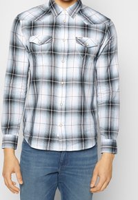 Camicia da uomo a maniche lunghe a quadri neri, bianchi e blu con colletto abbottonato, tasche sul petto e dettagli dorati. Tessuto di cotone, design aderente.