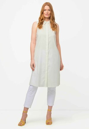 Lichtgroen mouwloos jurk met kraag, verticale knoopsluiting en een knie-lange snit, gecombineerd met witte cropped broek en gele sandalen met bandjes.