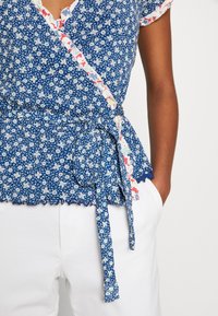 Haut cache-cœur bleu à motif floral avec bordure blanche et rose, manches courtes et ceinture à nouer. Ourlet festonné et tissu texturé ajoutent du détail.