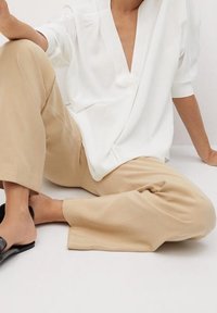 Blusa bianca, con scollo a V, drappeggiata in modo morbido, abbinata a pantaloni beige a gamba larga e sandali neri con apertura, mostrando un fit rilassato.