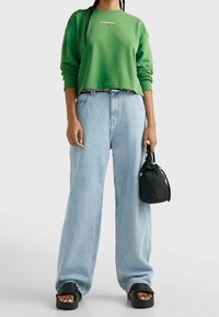Osoba v zelené mikině Tommy Jeans, světle modrých kalhotách s širokými nohavicemi, černých platformových sandálech, držící malé černé kabelky.