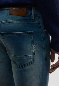 Φθαρμένα μπλε τζιν από τζιν με δερμάτινη ετικέτα, με έντονες ραφές σε αντίθεση στην τσέπη και κλασικό σχεδιασμό με πέντε τσέπες.