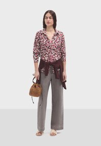 Camisa de botones con estampado floral en rojos y blancos, combinada con pantalones anchos grises. Sosteniendo un bolso trenzado marrón y vistiendo sandalias beige.