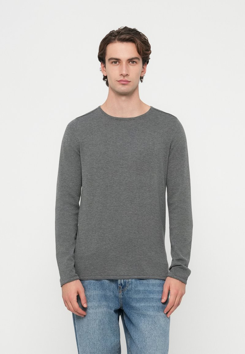 Siva majica z dolgimi rokavi, zaobljenim izrezom in mehko teksturo, kombinirana s svetlo modrimi jeans hlačami. Udoben in sproščen kroj.